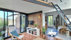 A spacious living area with a large window, a TV, a bookshelf, a blue chair, and a staircase. - L'Abri de la Plage, L'Abri de la plage - Maison de bord de mer en Bretagne (Lampaul-Plouarzel)