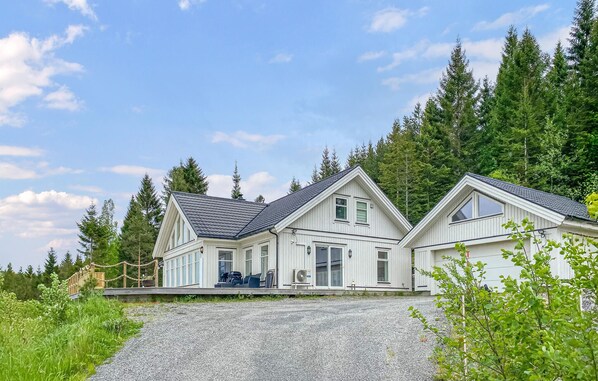 Une maison à deux étages avec une allée en gravier, entourée d’une forêt dense.