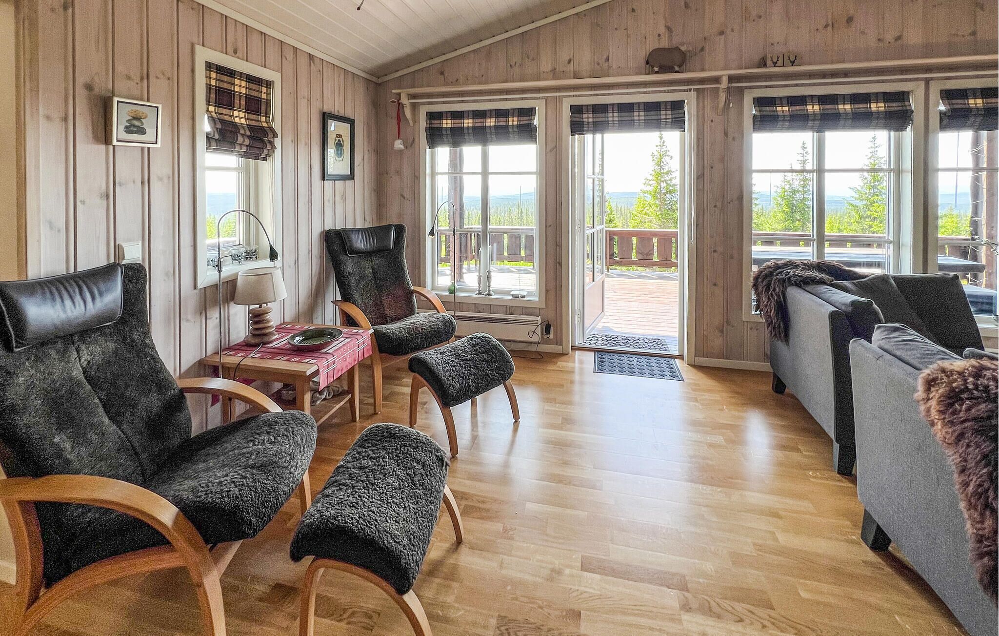 A cozy room with wooden walls, a wooden floor, a sofa, a chair, a table with a lamp, and a view through large windows.