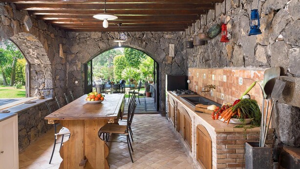 Una cucina esterna con un arco in pietra, tavolo in legno e angolo bar con utensili da cucina.