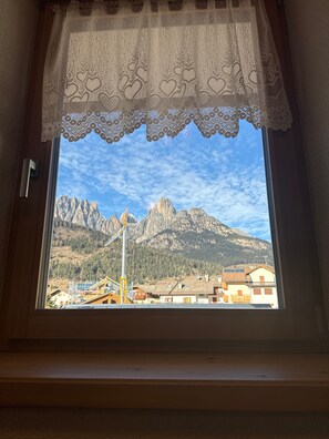 Une fenêtre ornée d’un rideau en dentelle, une vue sur les montagnes et des bâtiments au premier plan.