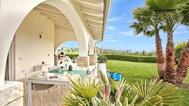 An outdoor patio with a table set for dining, surrounded by plants and palm trees. - Il Silenzio House- Manerba del Garda Villa With Garden (Manerba del Garda)