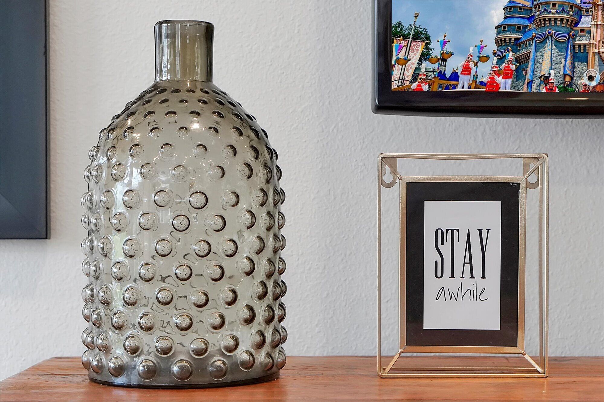 Eine dekorative Glasvase und ein gerahmtes Schild mit dem Schriftzug „STAY awhile“ auf einer Holztischfläche.