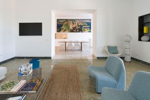 Un salon moderne avec une table basse en verre, un fauteuil bleu, un tapis à motifs et un coin repas comprenant une table et des chaises blanches.