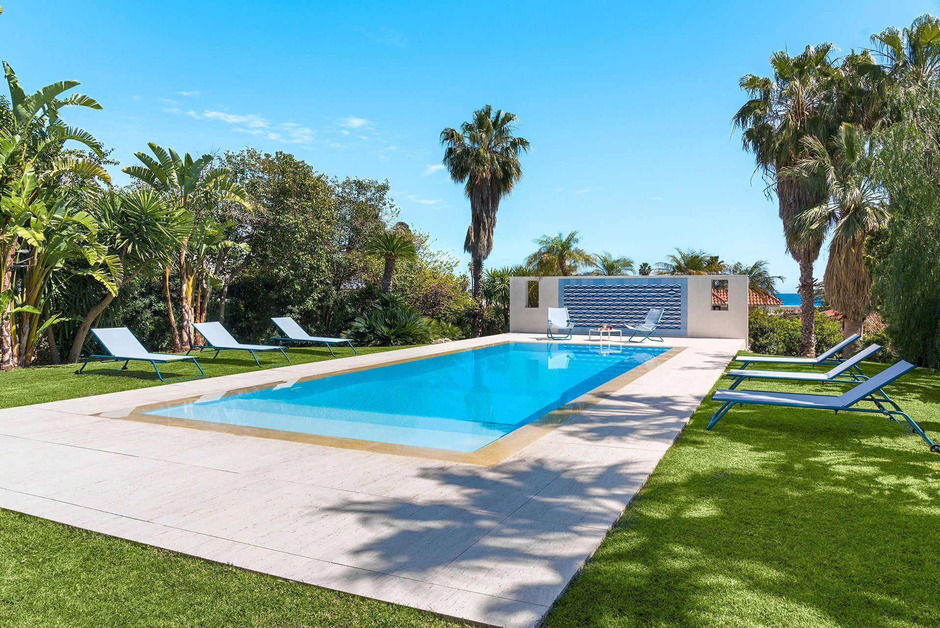 A rectangular swimming pool with a tiled deck, surrounded by green grass and palm trees.
