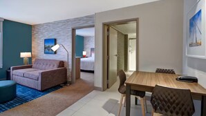 A hotel room with a sofa, a dining table with chairs, a lamp, and a bed in the background. - Spacious Room Near Entrust Bank Arena – Ideal for Sports Enthusiasts (Wichita)