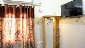 Een kamer met een wandgemonteerde flatscreen televisie, een airconditioning, een houten plank en een raam met een bloemenprint gordijn.