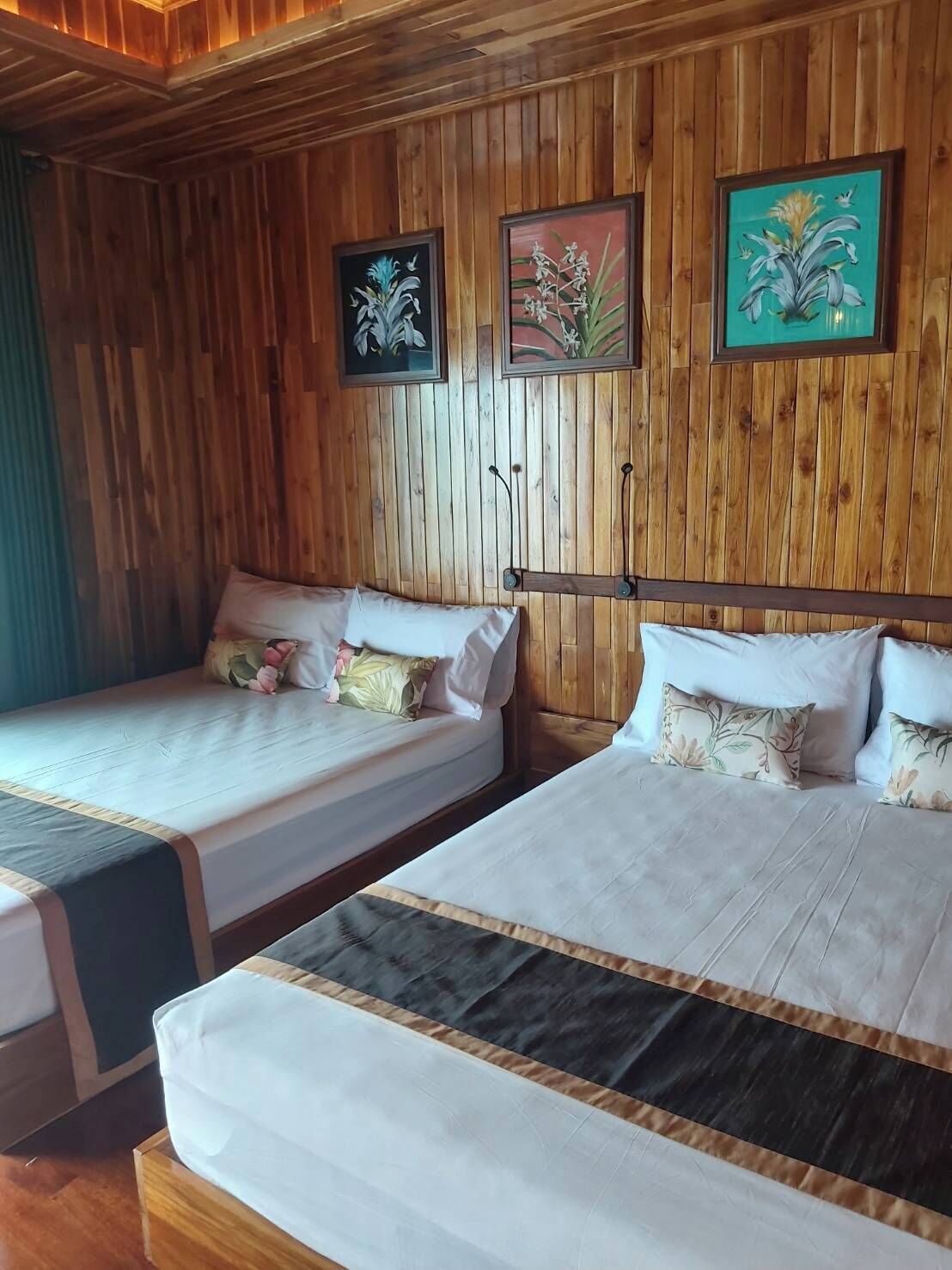 A room with wooden panel walls, two beds with white bedding, and three framed floral paintings.
