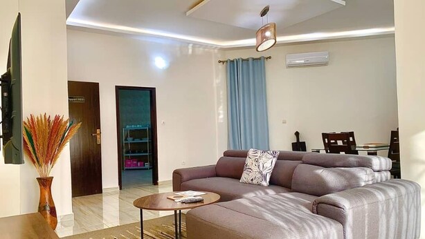 A modern living room with a sectional sofa, a coffee table, and a TV. There is a door leading to another room and a window with curtains. - HAIRAI-APPART HOTEL (Korhogo)