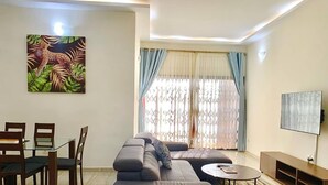 A modern living room with a sectional sofa, a dining table with chairs, a TV, and a large window with curtains. - HAIRAI-APPART HOTEL (Korhogo)