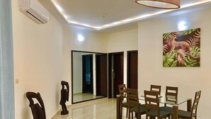 A dining area with a glass table and wooden chairs, a ceiling light fixture, and a wall-mounted air conditioner. - HAIRAI-APPART HOTEL (Korhogo)