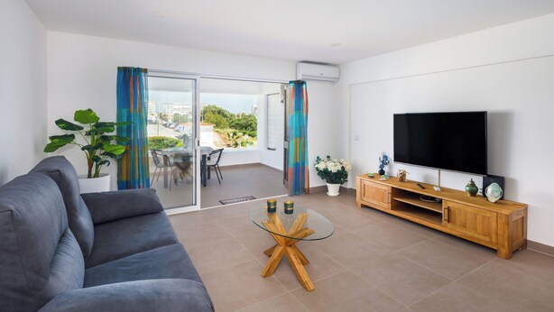 A modern living room with a grey sofa, a glass coffee table, a wooden TV stand, and a balcony with a view.