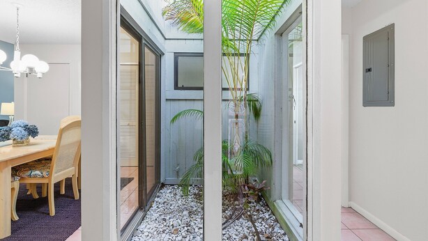 Un pasillo estrecho con una mesa y sillas de madera a la izquierda, un vidrio que da a un pequeño jardín con plantas, y una puerta de metal a la derecha.