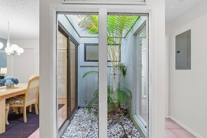 Ein schmaler Flur mit einem Holzesstisch und Stühlen links, einer Glastür, die zu einem kleinen Garten mit Pflanzen führt, und einer Metalttür rechts.