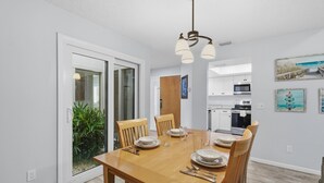 Un coin repas avec une table en bois dressée pour quatre personnes, une cuisine en arrière-plan et une porte-fenêtre coulissante donnant sur un balcon agrémenté de plantes.
