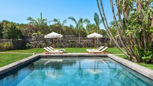 A rectangular swimming pool with clear blue water, surrounded by a stone wall and lush greenery. There are two lounge chairs with umbrellas and a stone pathway leading to the pool.