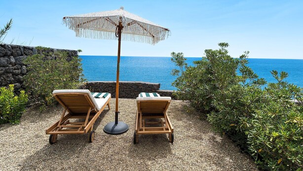Two wooden lounge chairs with striped cushions, a woven parasol, and a view of the sea.