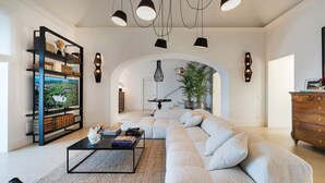 A modern living room with a sectional sofa, a coffee table, and a bookshelf. There is a staircase leading to an upper level, and a large window providing natural light.