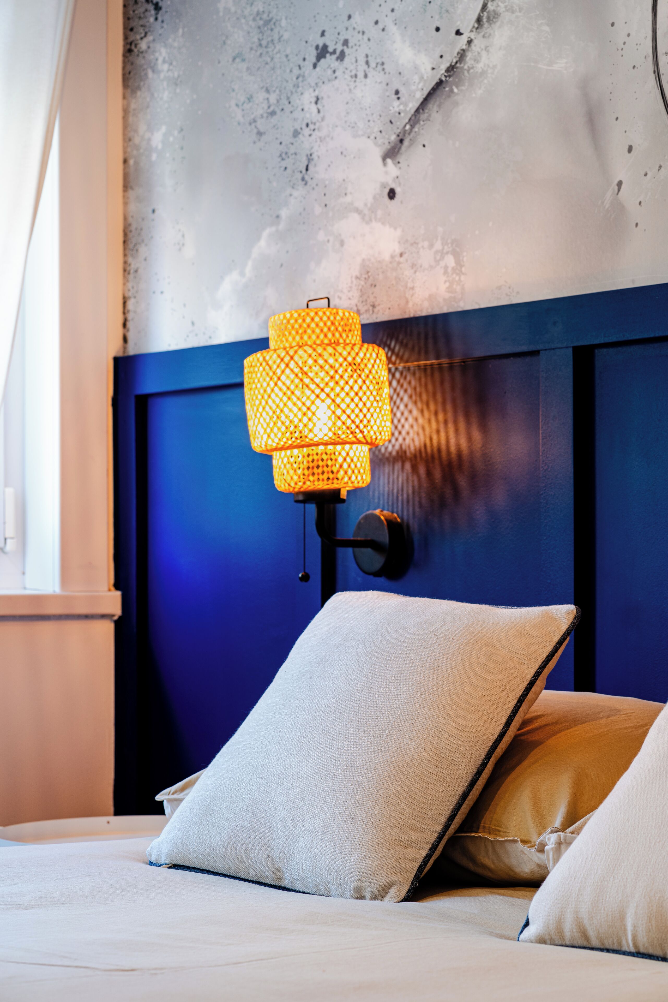 A bed with white bedding and two beige pillows, a wall-mounted lamp with a yellow patterned shade, and a blue headboard.