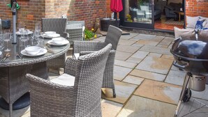 Een terras buiten met rieten meubilair, een barbecue en een rode parasol.