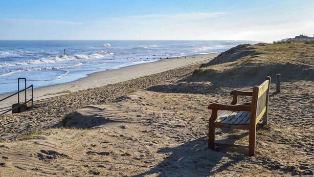 Een strand met een houten bankje en een wachthuisje in de verte.