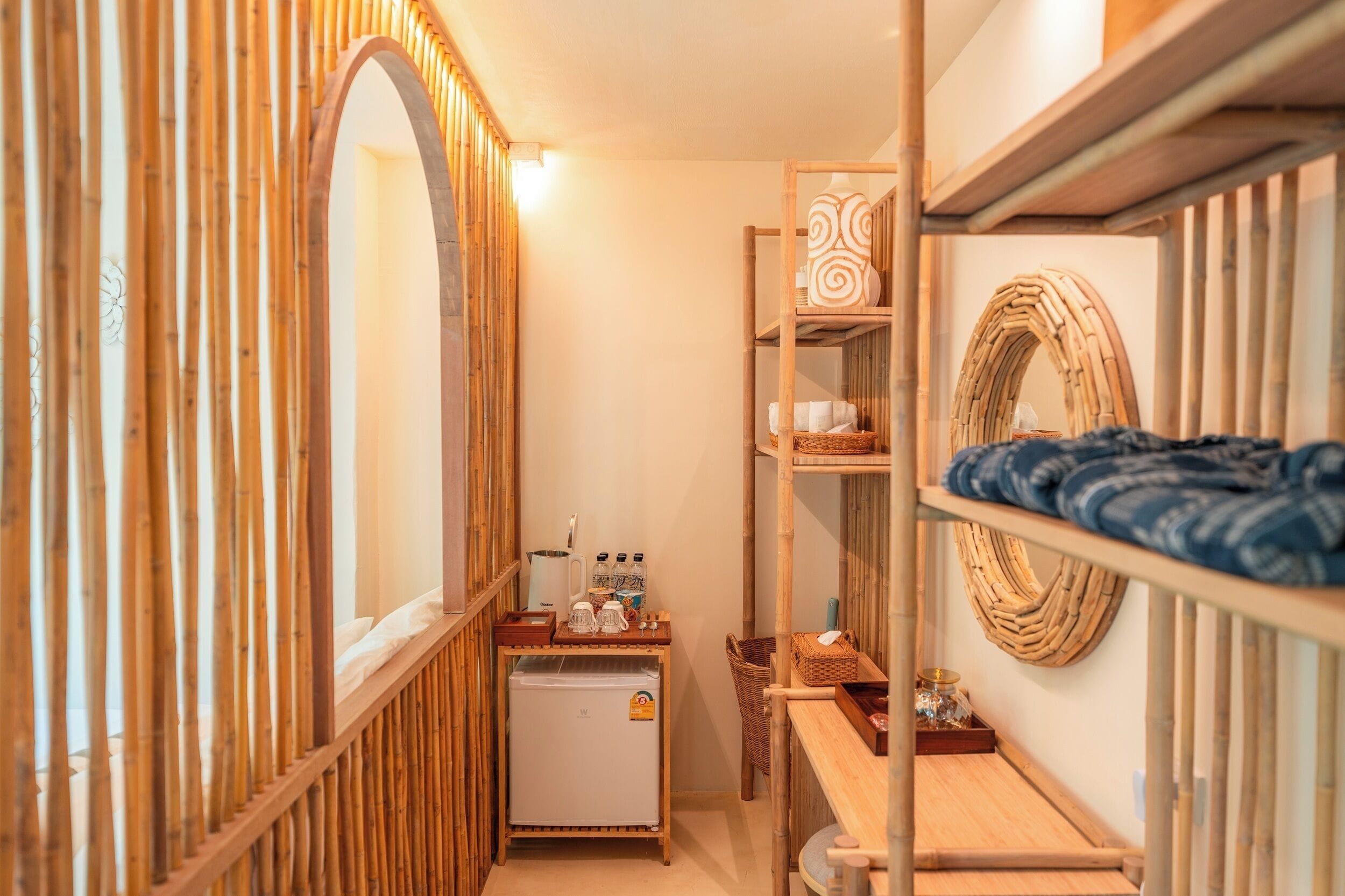 A compact room with a bunk bed, a small refrigerator, a shelf with decorative items, and a mirror.