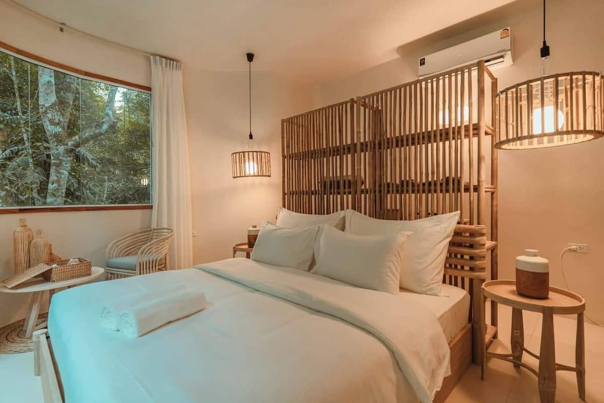 A modern bedroom with a large bed, a wooden headboard, and a view of trees through a window.