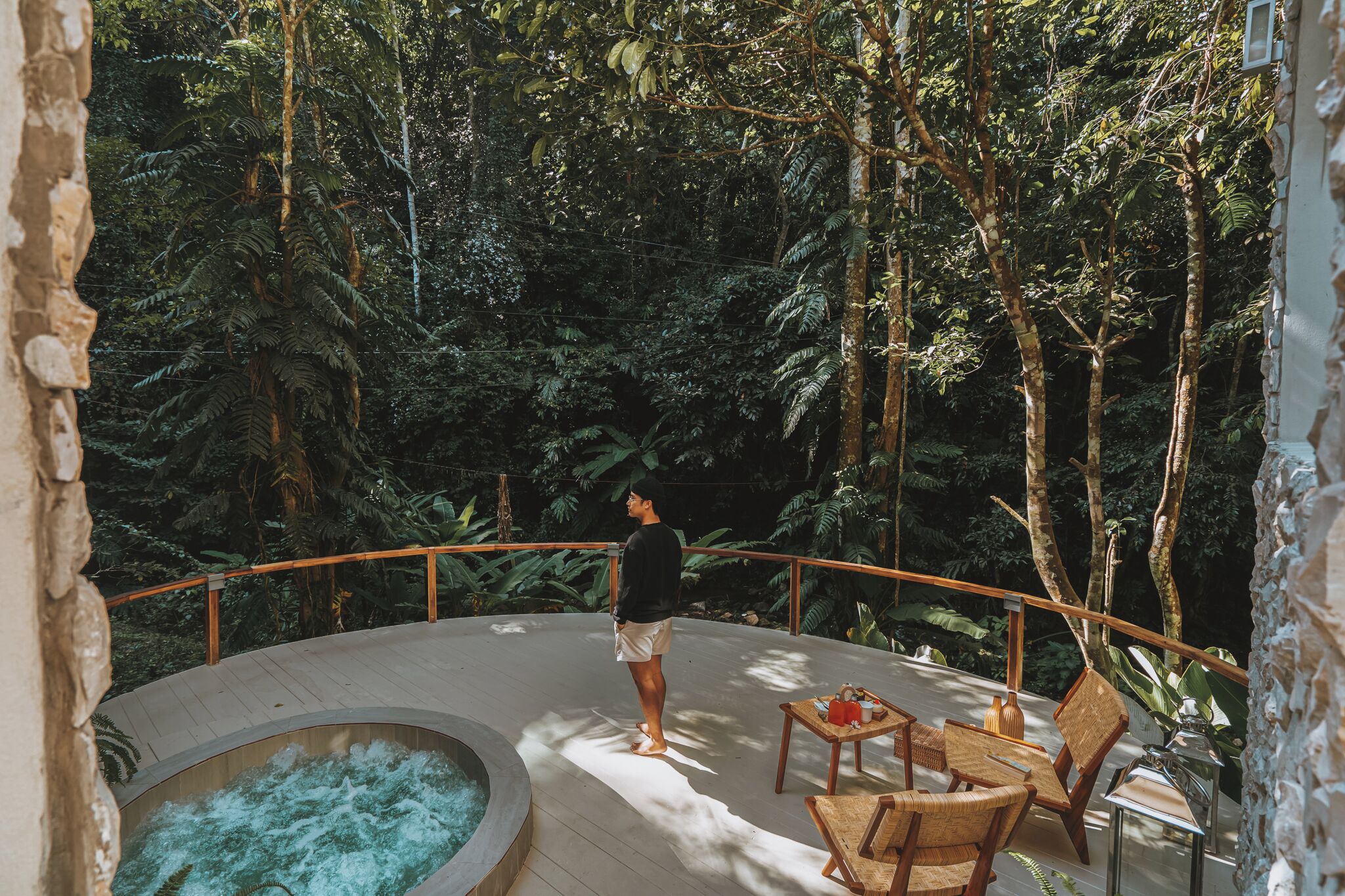Un hombre está de pie en una terraza con vistas a un jacuzzi y abundante vegetación.
