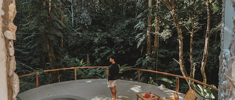 Un hombre está de pie en una terraza con vistas a un jacuzzi y abundante vegetación.