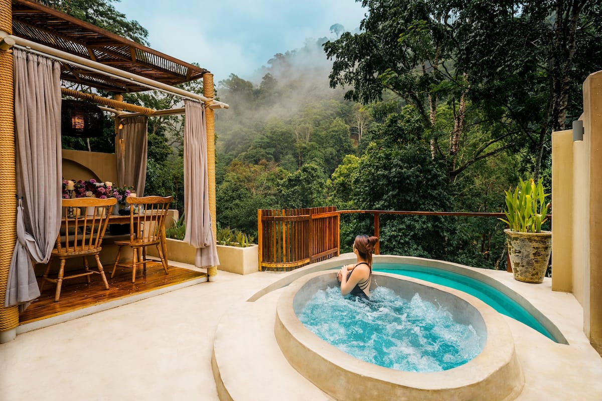 A hot tub with a view of a lush forest, a wooden deck with chairs, and a pergola with curtains.