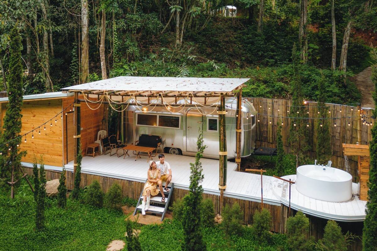 A wooden RV with a covered porch, a hot tub, and a deck area surrounded by trees.