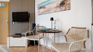 A room with a TV, a painting of a beach, a small table with a lamp, and wicker chairs.