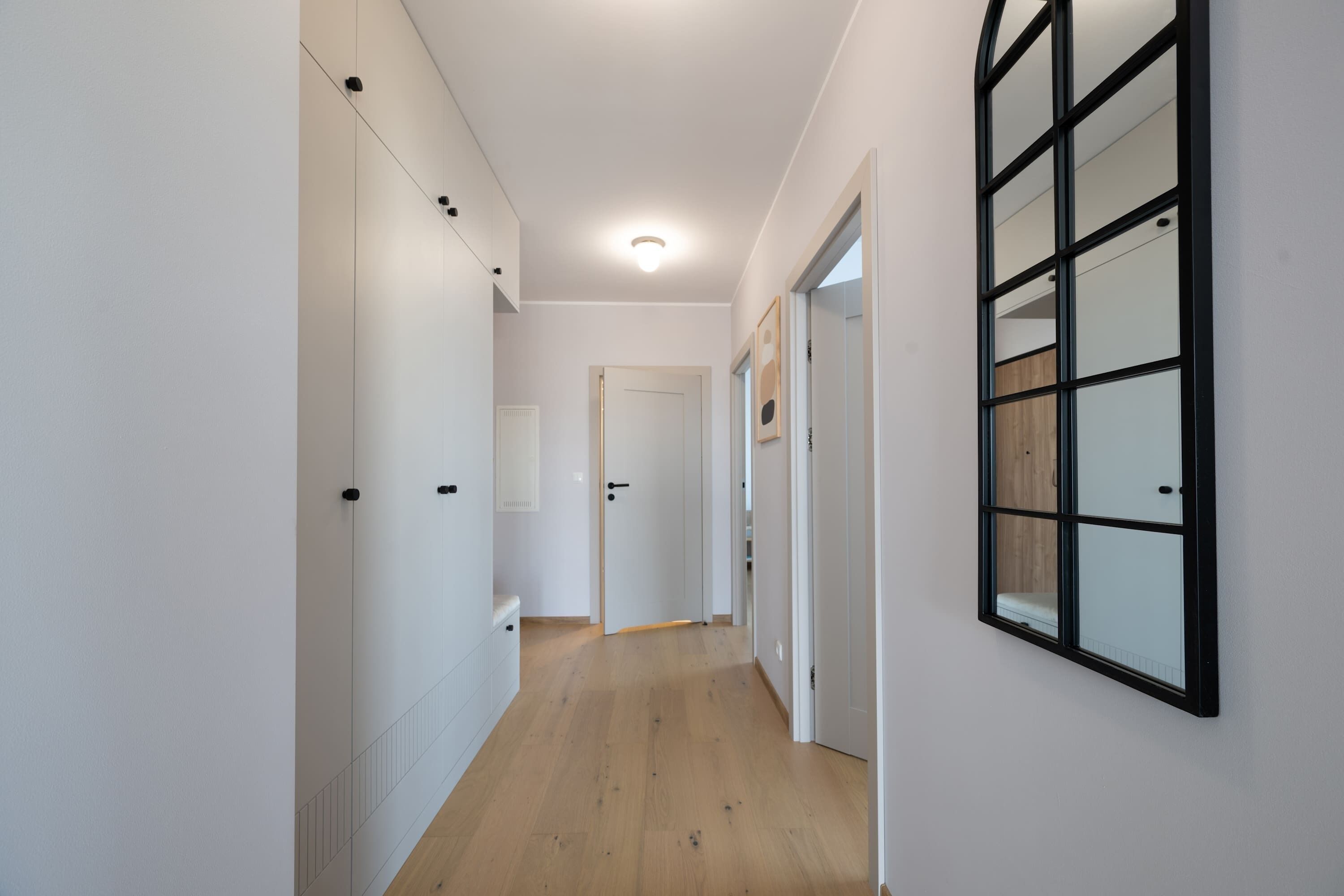 A narrow hallway with wooden flooring, white walls, and a large window with multiple panes.