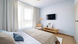 Una camera da letto con un letto, una TV a parete, una sedia, un comodino con una pianta e una finestra con tende.