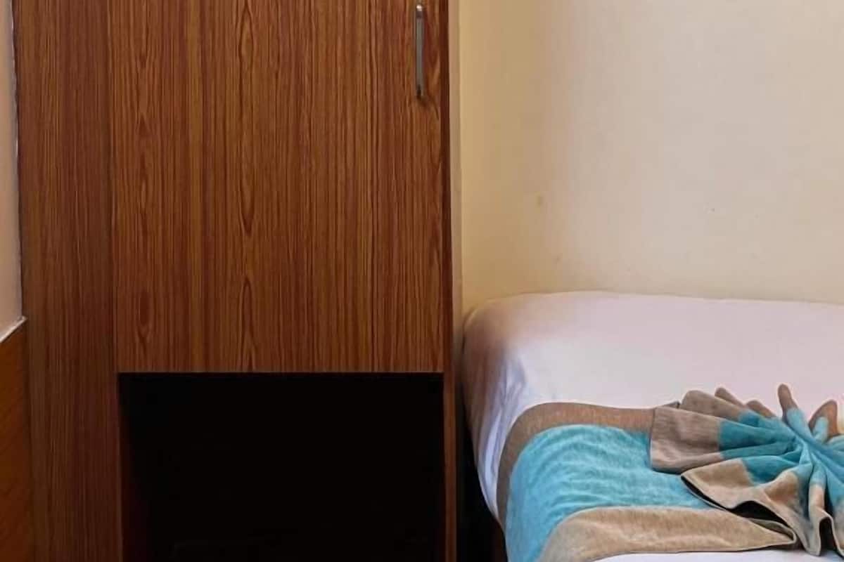 A room with a wooden wardrobe, a bed with a blue and beige blanket, and a white wall.
