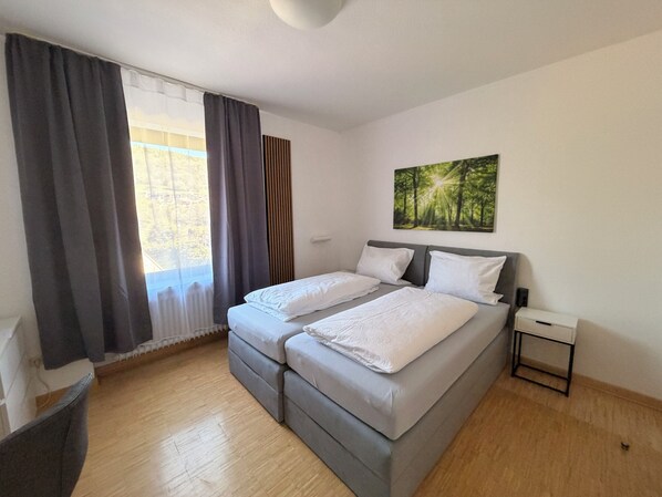 A bedroom with a bed, a nightstand, a window with curtains, and a painting on the wall. - Klosterhof Neustadt am Main (Neustadt a. Main)