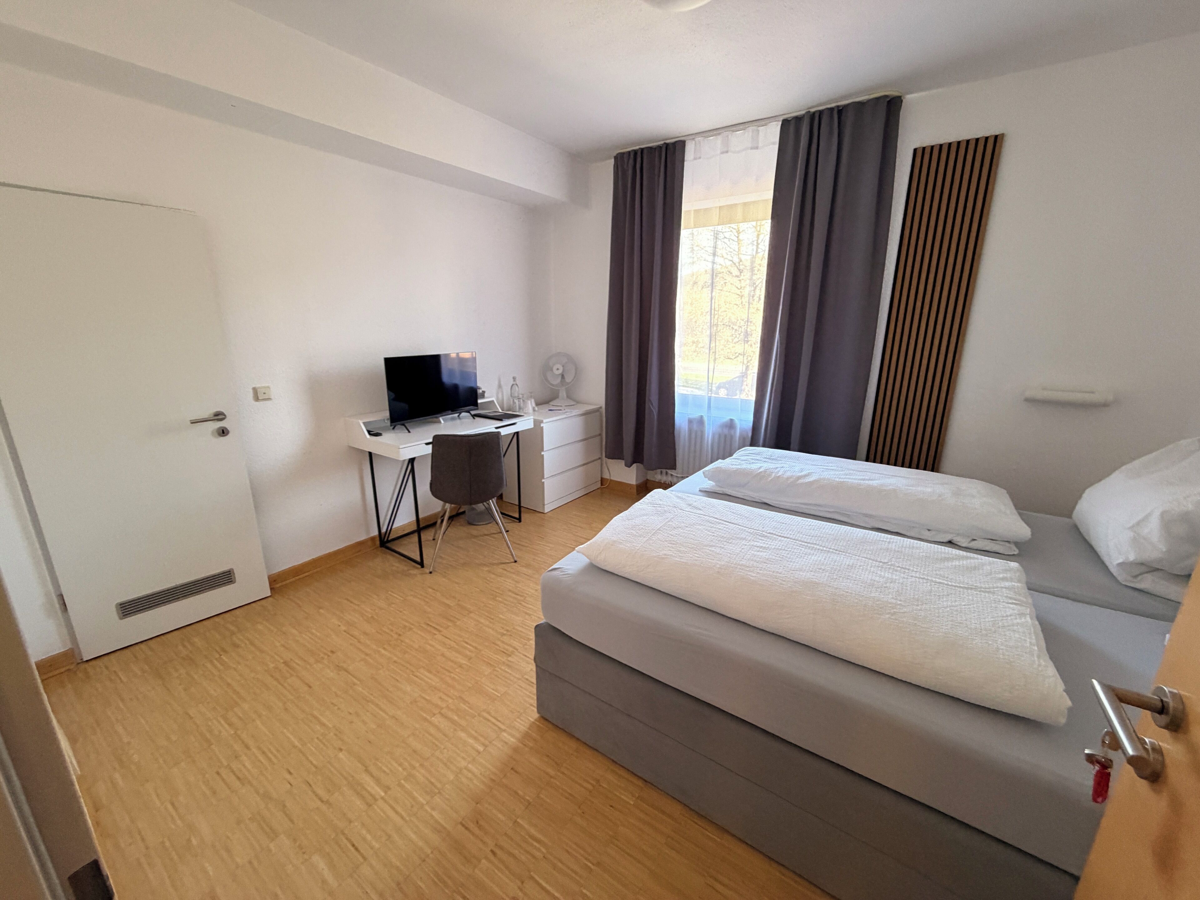 A hotel room with a bed, a desk with a computer, a chair, and a window with curtains.