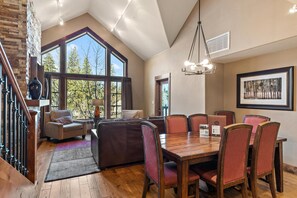 A spacious dining area with a high ceiling, large windows, and a view of trees outside. - Riversong | Exclusive Tamarack Perks (Tamarack)