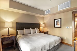 Une chambre d’hôtel avec un grand lit, deux tables de chevet, une petite table, un tableau encadré au mur et une fenêtre avec des stores.