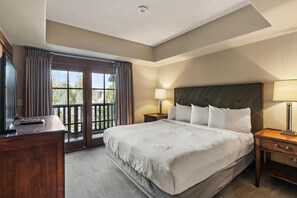 A hotel room with a large bed, a desk, a chair, a nightstand, a lamp, and a view of the outdoors through a window. - Riversong | Exclusive Tamarack Perks (Tamarack)