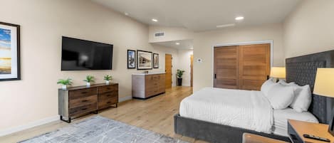 A spacious bedroom with a large bed, a TV mounted on the wall, a wooden wardrobe, and a desk with a lamp.