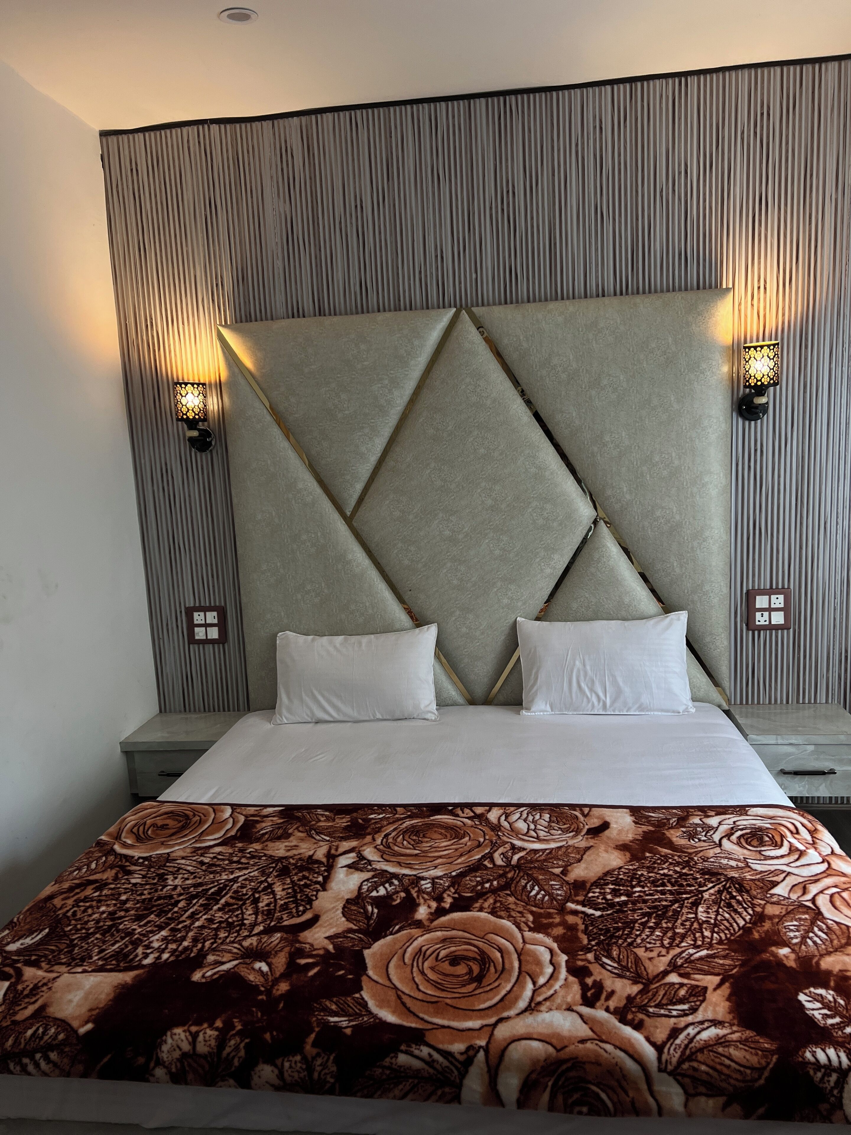 A hotel room with a large bed, two pillows, a decorative headboard, two wall-mounted lamps, and a nightstand.