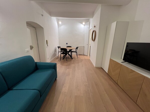 A modern living room with a teal sofa, a dining area with a table and chairs, and a TV on a wooden cabinet.