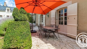 A patio with a table and chairs under an orange umbrella, a white door, and a stone-tiled floor. - Elegant B&B in La Ménitré with heated pool and wifi (La Ménitré)