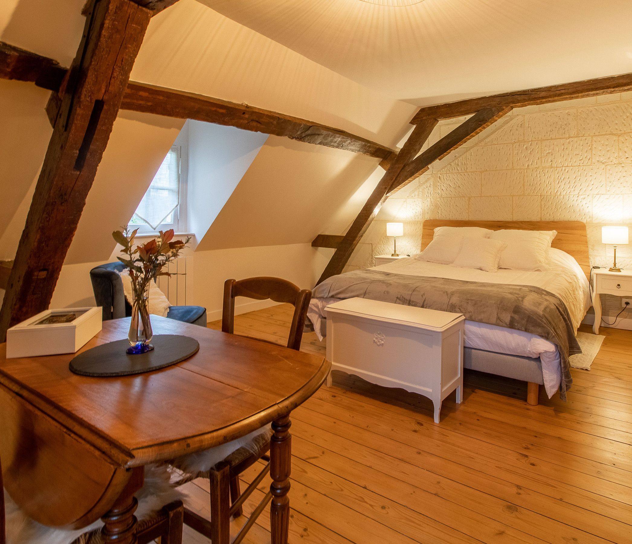 Um quarto de sótão acolhedor, com teto de madeira, uma cama com roupa de cama branca, uma mesa de cabeceira com abajur e uma pequena mesa com um vaso de flores.