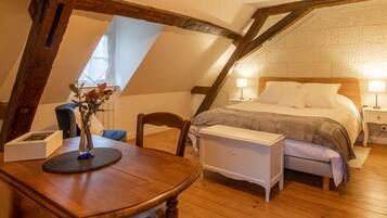 Une chambre confortable située sous les toits, avec un plafond en bois, un lit recouvert de draps blancs, une table de chevet avec une lampe et une petite table sur laquelle est posé un vase de fleurs.