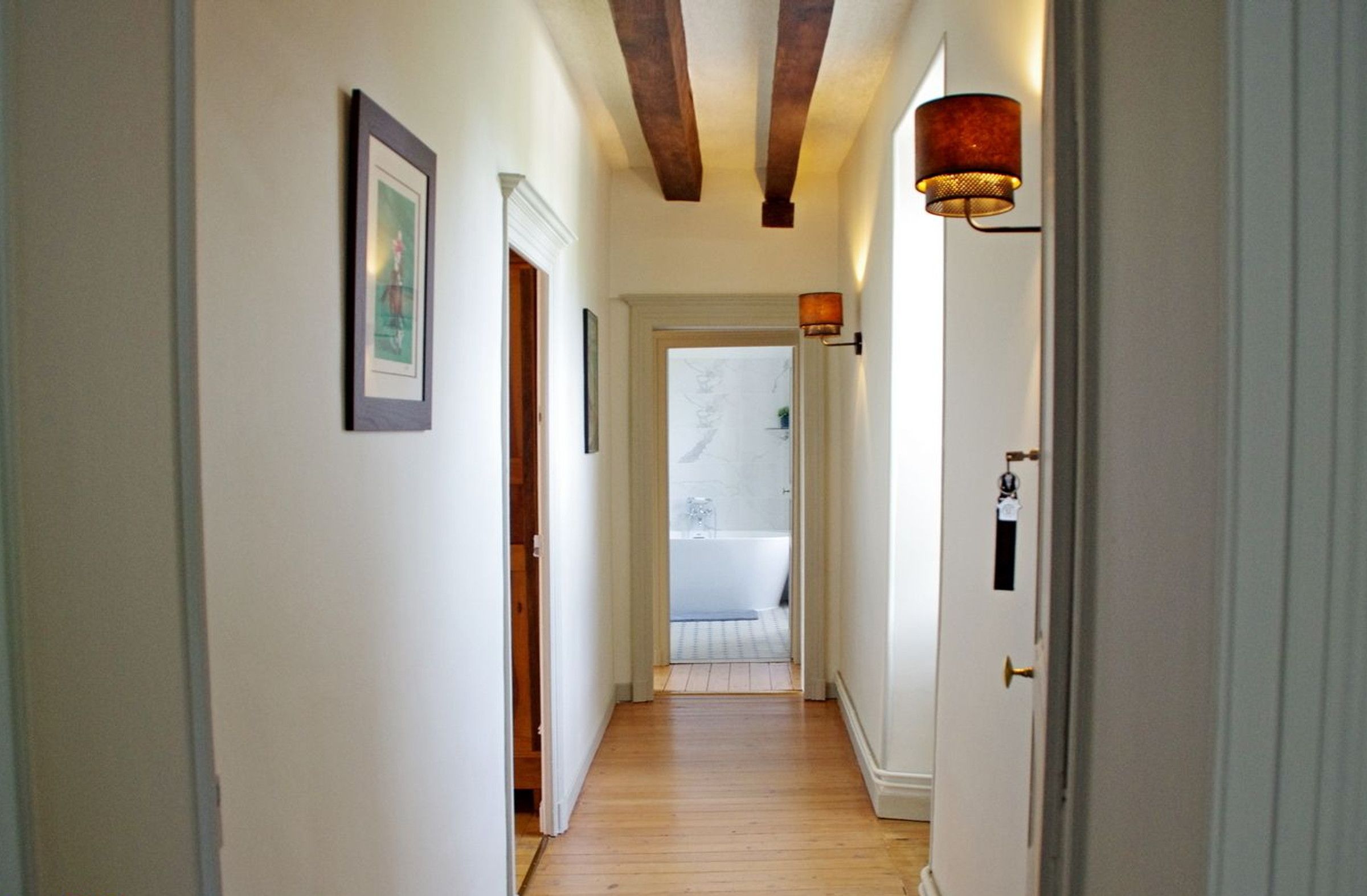 Un couloir doté d’un plancher en bois, de murs blancs et d’une porte donnant sur une salle de bain avec une baignoire.
