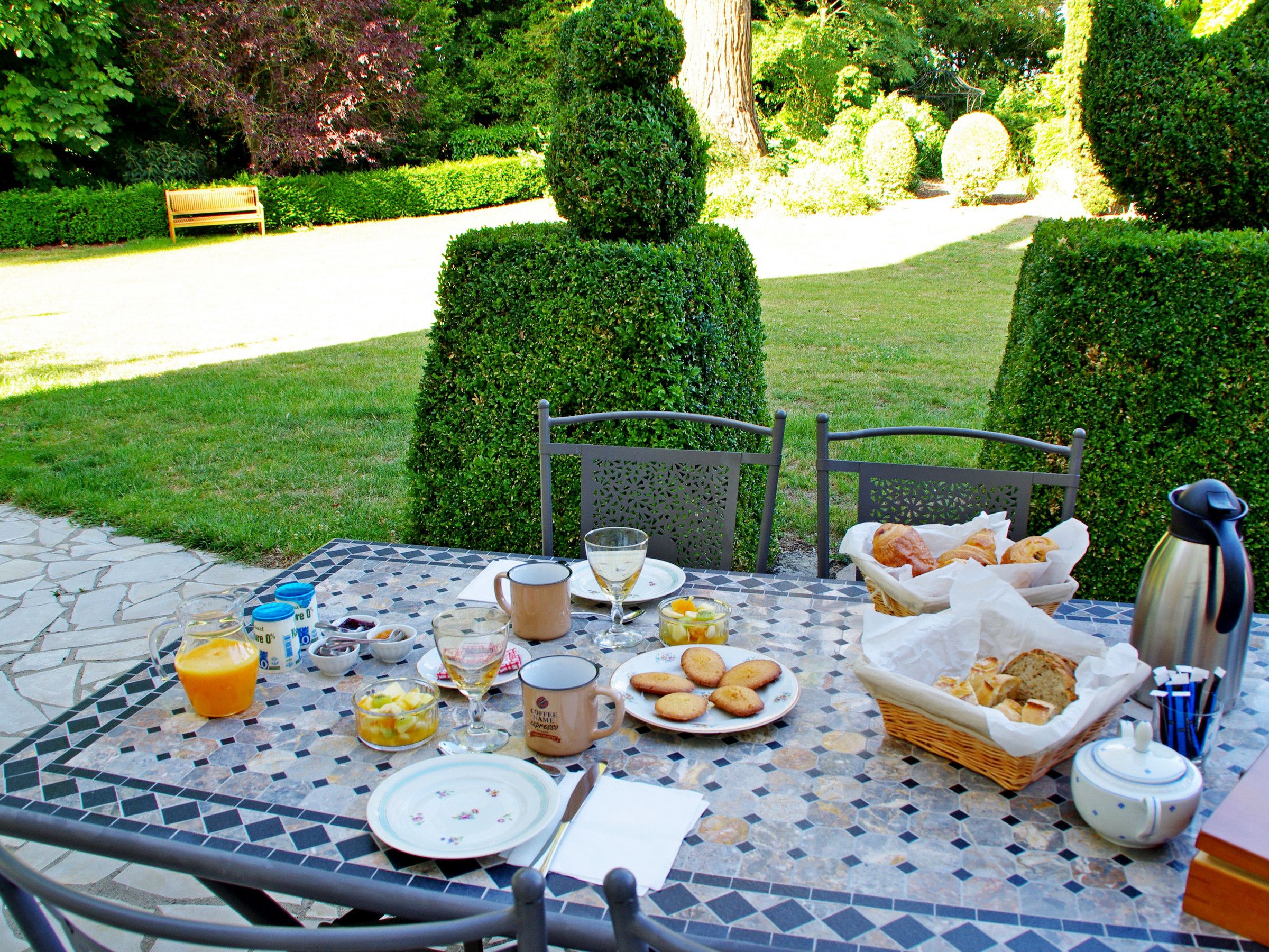 Un tavolo da pranzo all'aperto con cibi per la colazione, una panca e siepi curate sullo sfondo.