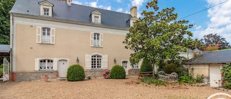 A two-story house with a beige facade, multiple windows with shutters, and a gravel driveway.