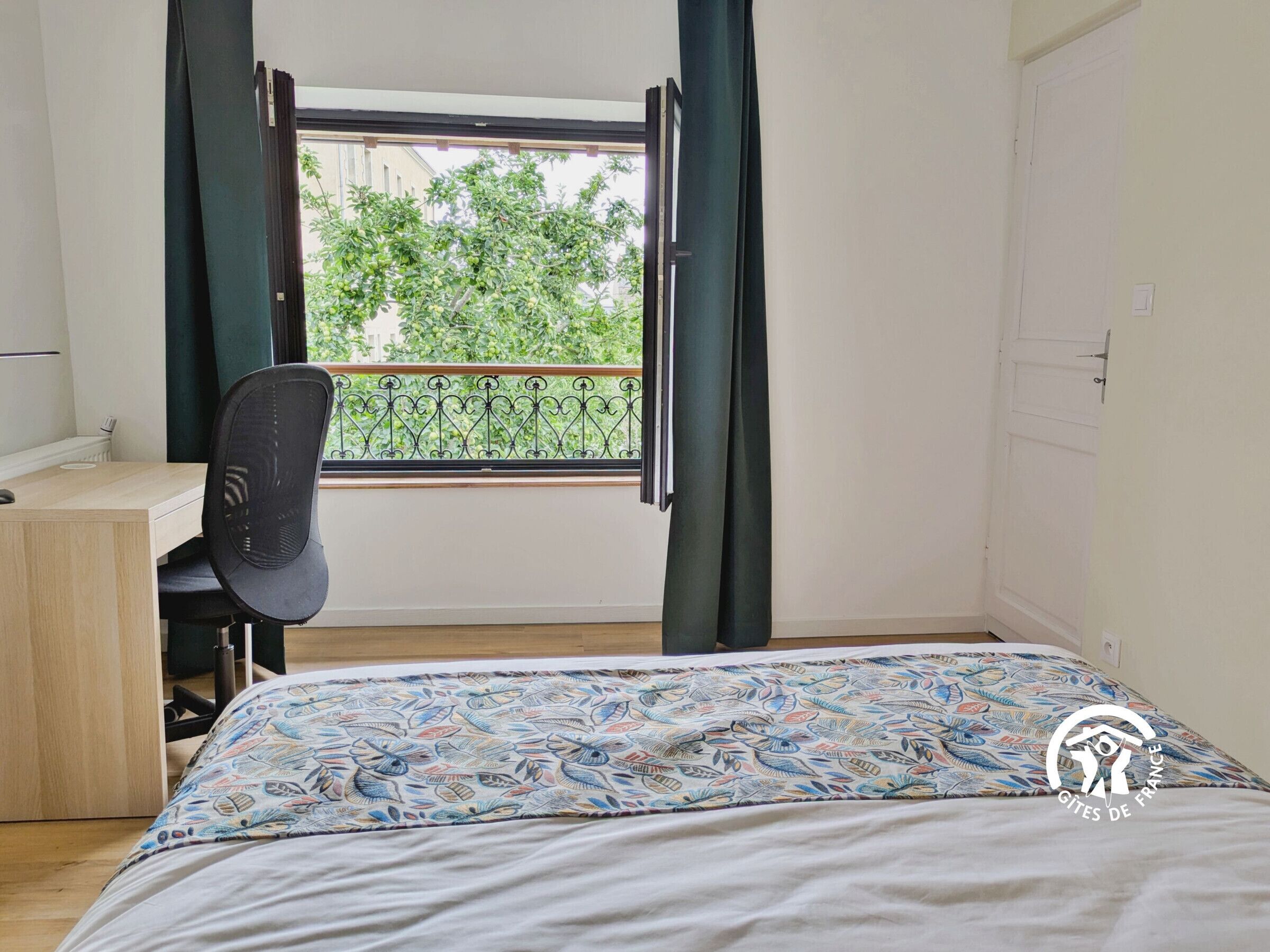 A bedroom with a bed, a desk, and a chair. There is a window with curtains and a view of greenery outside.